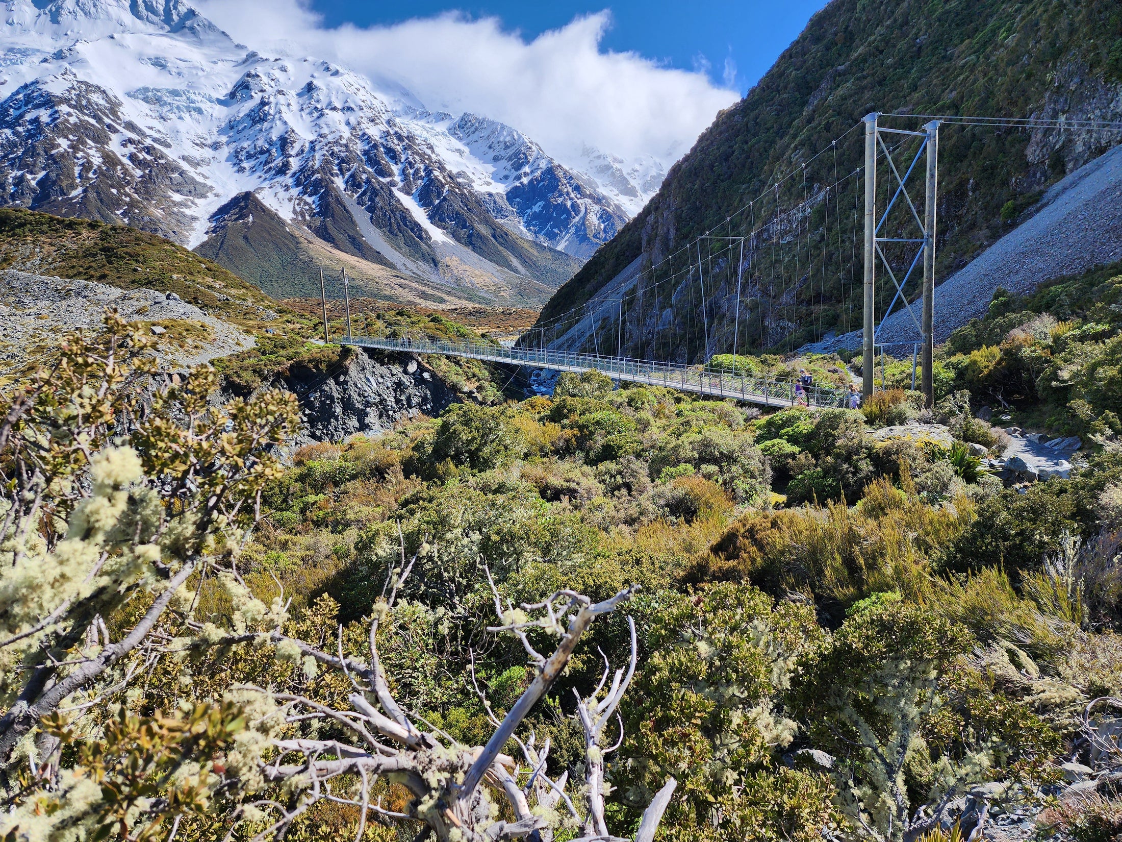 A scenic view of a landscape with mountains, a river, and a suspension bridge, with a part of the mountain covered in snow.