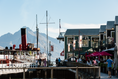 Load image into Gallery viewer, A view of Queenstown's waterfront with a mountain in the background, featuring a clear blue sky.
