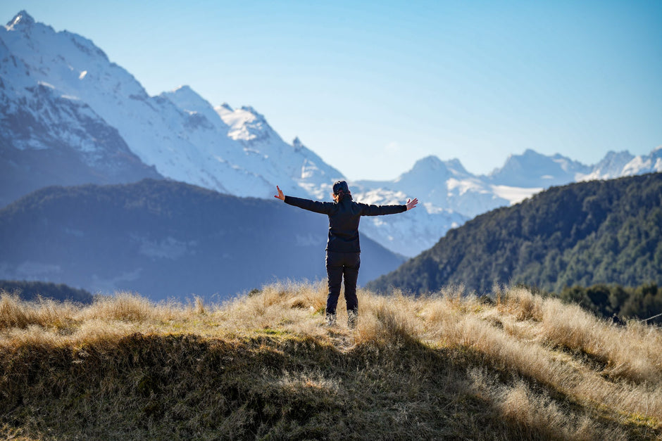 A walker with arms outstretched on a tussock ridge, facing the snow-covered peaks at the head of Lake Wakatipu