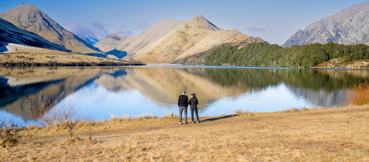 Two walkers on the shore of Moke Lake, with the tussock hills mirrored in the water